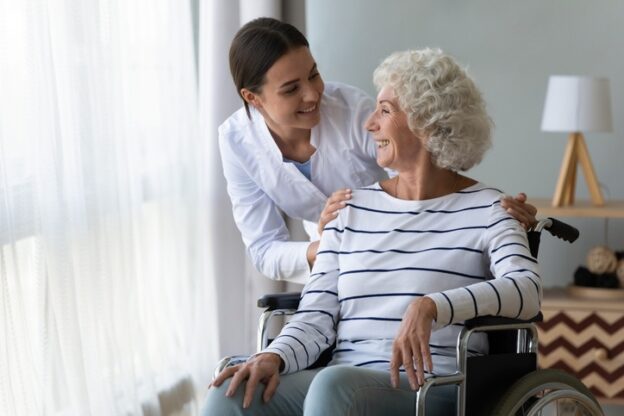 Smiling disabled elderly woman sitting in wheelchair talking with caring young nurse in living room
