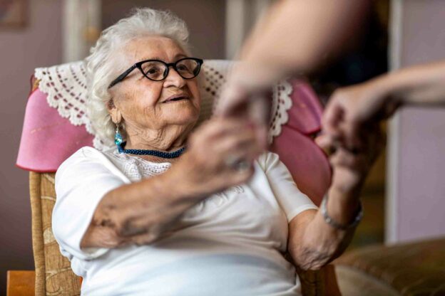 An older woman sitting in a chair reaching out her hands to another person