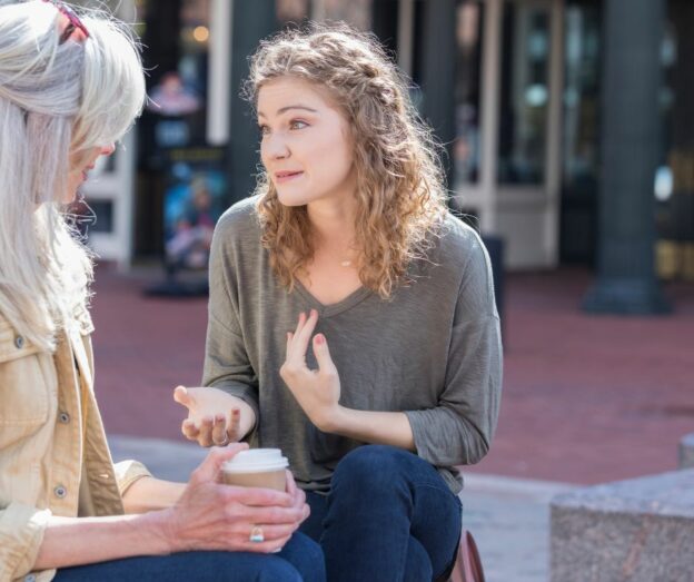 Two women speaking sat in the middle of the street, one is drinking a coffee
